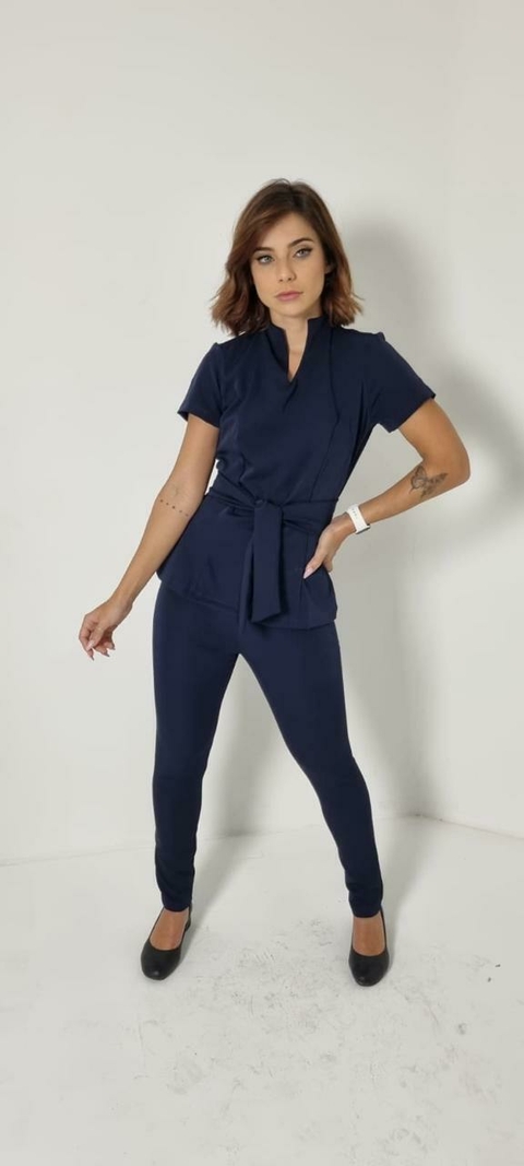 Mulher vestindo scrub azul marinho Cíntia Flex com calça justa, blusa com gola diferenciada e faixa na cintura, em fundo branco
