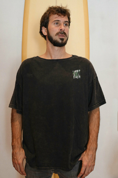 Hombre usando un remerón negro con un diseño en el pecho, de pie frente a una tabla de surf.