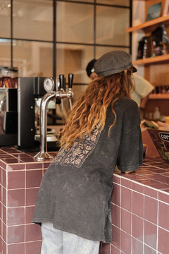 Mujer de espaldas usando un remerón negro con diseño de mosaico, apoyada en la barra de un café.