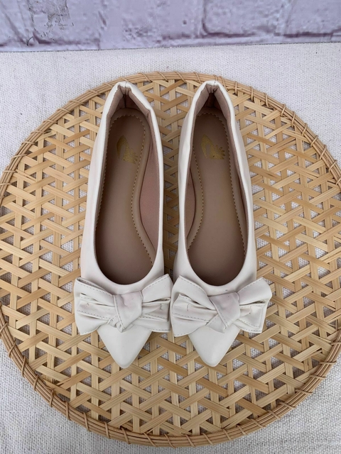 Sapatilhas alongadas off-white com laço nó na parte frontal, exibidas em cima de um suporte de palha circular