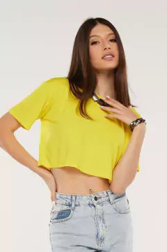 Modelo morena usando um cropped liso feminino amarelo em viscolycra, sorrindo e olhando para a câmera, mão direita na cintura, mão esquerda proxima do pescoço, sobre fundo branco. G&B Bros