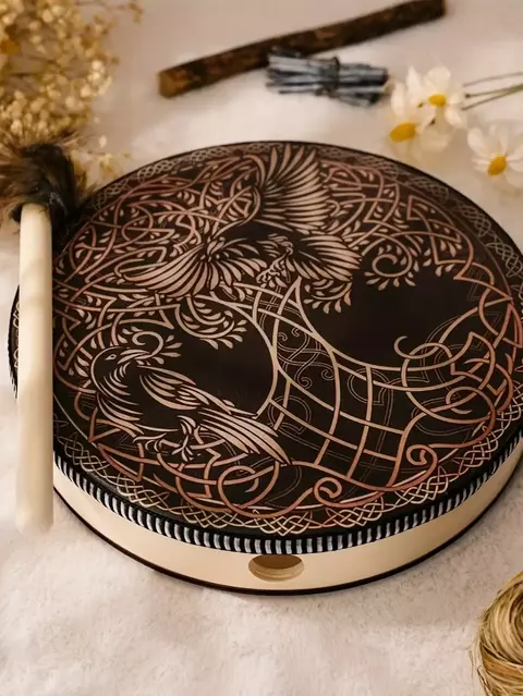 Tambor xamânico vegano decorado com desenhos intricados, colocado sobre uma superfície clara, ao lado de um bastão de percussão e flores.