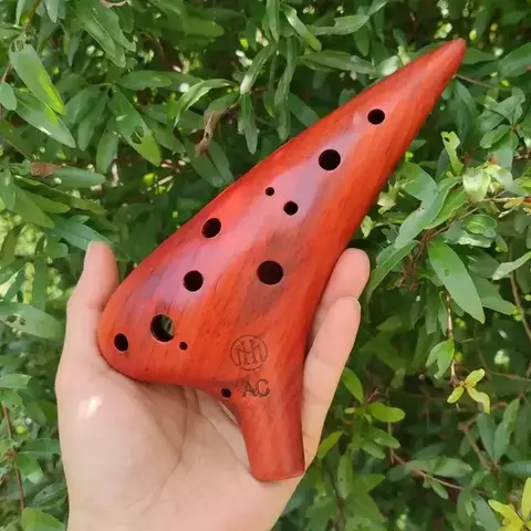 Ocarina em bambu de 12 buracos, segurada por uma mão, com fundo de folhas verdes.