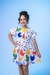Menina sorridente usando um vestido branco com estampas coloridas de pássaros e flores.