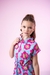 Menina usando um macaquinho azul com estampa de flores coloridas em fundo rosa.