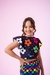 Menina sorrindo vestindo um conjunto colorido com estampas florais e xadrez, posando em um fundo rosa.