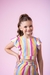 Menina sorrindo usando um conjunto listrado colorido, composto por blusa e saia, com fundo rosa.