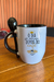 Caneca com a frase "O dia só começa depois do café" e colher preta, sobre uma mesa de madeira.