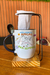 Caneca com a frase "Melhores Amigas Pra Sempre", acompanhada de uma colher e um suporte com pano, sobre uma tábua de madeira.