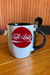 Caneca branca com estampa vermelha "Café com Leite" e colher ao lado, sobre uma mesa de madeira.