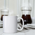 Caneca reta branca de porcelana com capacidade de 150ml, exibida em superfície clara com utensílios de mesa ao fundo