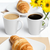 Canecas cônicas brancas de porcelana de 150ml com café e café com leite, acompanhadas de croissants em pratos brancos sobre mesa posta com flores amarelas