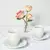 Xícaras de chá cônicas de porcelana branca com pires, acompanhadas por vaso pequeno com três rosas cor de rosa e amarelas.