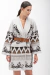 Imagem de um cardigan branco com detalhes em preto e marrom, com padrão geométrico e floral, sendo usado por uma mulher com cabelo liso preso, usando um cinto marrom na cintura.