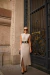 Mulher usando um vestido envelope de nervuras na cor bege, com fenda, cinto preto e acessórios elegantes, posando em frente a uma porta decorativa.