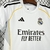 Camisa infantil do Real Madrid 2025/26, predominantemente branca, com detalhes em preto e amarelo, e logotipo do clube.