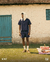 Modelo masculino usando uma camisa boxy xadrez azul, calça curta e tênis, em um cenário ao ar livre com um fundo de parede de tijolos e uma porta azul.