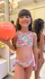 Biquíni infanto juvenil cropped kids na cor rosa, com estampa colorida de escamas, usado por uma menina sorridente segurando uma bola laranja.