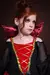 Criança vestida com fantasia de vampira luxuosa, com cabelo ruivo e vestido preto e vermelho, detalhes em dourado.