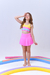 Menina usando uma saia de praia infantil curta na cor rosa, com um top amarelo e detalhes em rosa e azul, fazendo uma pose divertida.