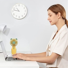 Una mujer utilizando un ventilador de mano con respaldo angular mientras trabaja en su computadora portátil, con un reloj de pared en el fondo.