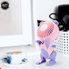 Ventilador de escritorio en forma de animal, color rosa y morado, con soporte para celular, ubicado sobre una mesa junto a una taza de café.