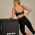 Top Rafa feminino de treino com alças finas e recorte cruzado, ideal para exercícios intensos. Disponível nas cores preto e marrom.