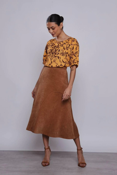 Mulher vestindo saia midi evasê de veludo cotelê castanho, combinada com blusa estampada amarela.