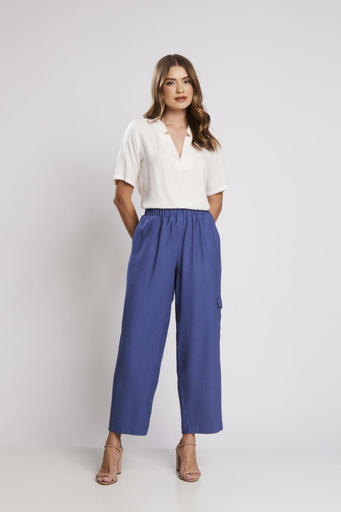 Mulher vestindo calça azul com elástico na cintura, combinada com blusa branca de manga curta e sandálias de salto, em fundo neutro.