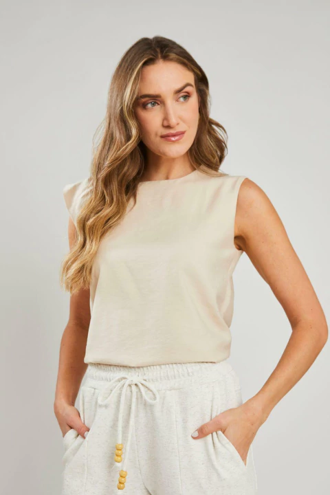 Mulher vestindo uma blusa regata assimétrica bege, modelo Helena, com calça clara, em fundo neutro. Peça feminina elegante e casual, ideal para o verão.