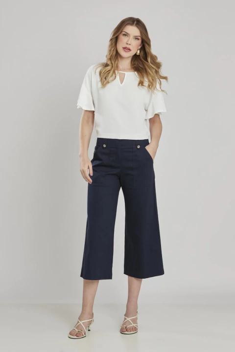 Mulher vestindo calça cropped reta na cor marinho, combinada com blusa branca de manga curta e sandálias claras, em fundo neutro.