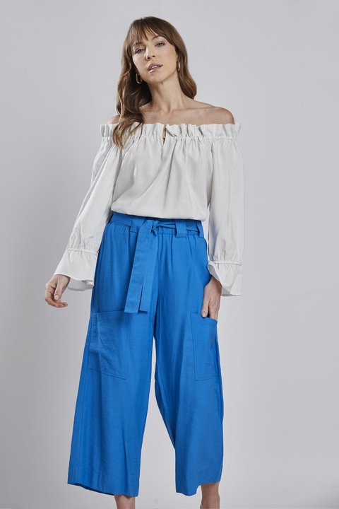 Mulher vestindo blusa franzida ombro única branca e calça azul, posando para foto de moda feminina.