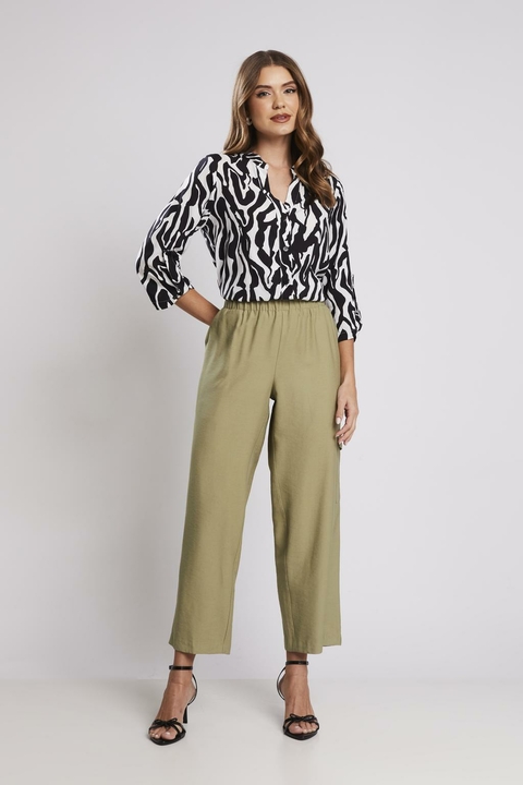 Mulher vestindo calça com elástico verde folha, combinada com camisa estampada preta e branca e sandálias de salto, em fundo neutro.