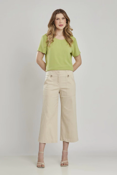 Mulher vestindo calça cropped reta na cor areia, com blusa verde clara e sandálias de salto, em fundo neutro. Moda feminina elegante e ideal para office look.