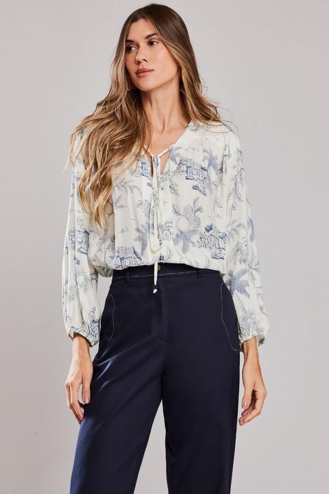 Mulher vestindo blusa bata Porto Unica estampada de verão, com mangas longas e calça azul escura, em fundo neutro.