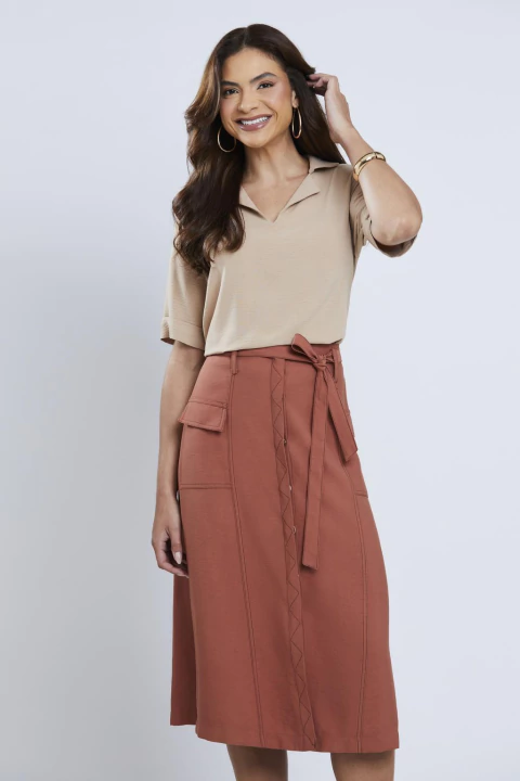 Mulher vestindo blusa bege de manga 3/4 com gola V e saia midi terracota, posando sorridente em fundo neutro.