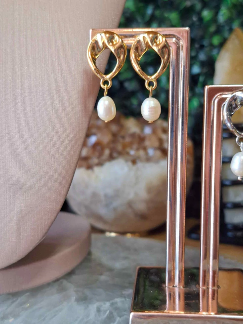 Brincos dourados com design orgânico e pérolas de água doce penduradas, exibidos em suporte metálico