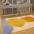 Tapete infantil Kamet com design abstrato em cores amarelo, laranja, azul e branco, posicionado em um quarto com móveis modernos.