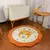 Tapete infantil orgânico redondo com desenho de girafa sorridente em tons de laranja e branco, colocado no chão de um quarto com berço e mesinha de madeira