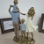 Escultura em resina representando uma família com pais e filhos, em tons dourados e com a mãe vestindo uma camisa azul e uma saia branca.