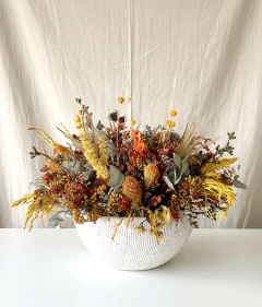 Arranjo seco de flores em vaso redondo de cerâmica branca com flores e folhagens em tons amarelo, laranja, marrom e verde