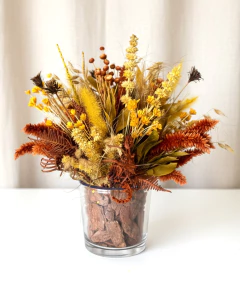 Arranjo seco em tons de amarelo, laranja  e marrom em vaso de vidro transparente com casca de pinus