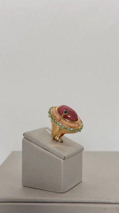 Anel dourado ajustável com design arredondado, com pedra central rosa, cristais verdes e detalhes em cristal azul