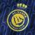 Escudo do Al-Nassr estampado no lado esquerdo do peito da camisa Away Reserva 2024/2025 Adidas azul marinho masculina, com padrão gráfico em azul royal, usada por Cristiano Ronaldo, CR7, Anderson Talisca e Mané.