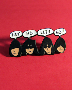 Pin de metal com desenho do rosto dos integrantes da banda Ramones dizendo "hey ho let's go!". Possui dois fechos na parte de trás.