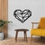 Quadro decorativo em forma de coração, feito em madeira, com design vazado e moderno, instalado em uma parede clara ao lado de uma mesa redonda e uma cadeira amarelo-ocre.