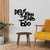 Quadro decorativo em MDF com a frase "Deus é Bom o Tempo Todo", exibido em um ambiente moderno com mesa e sofá amarelo.