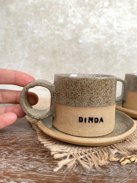 Xícara de cerâmica com a palavra "DINDA" gravada, acompanhada de pires da mesma cor