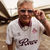 An older man wearing a cream and burgundy Köln Retrô 2025 jersey, sunglasses, and a silver chain, standing outdoors.