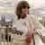 Woman wearing a cream and burgundy Köln Retrô 2025 jersey with sunglasses, standing on a balcony with a cityscape background.